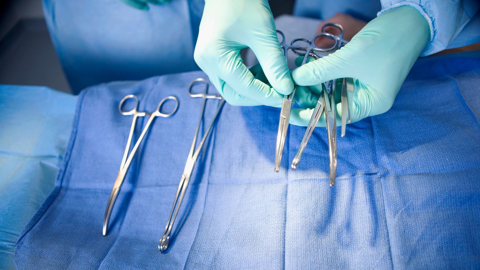 Two gloved hands holding surgical tools over a table covered in a blue sheet.