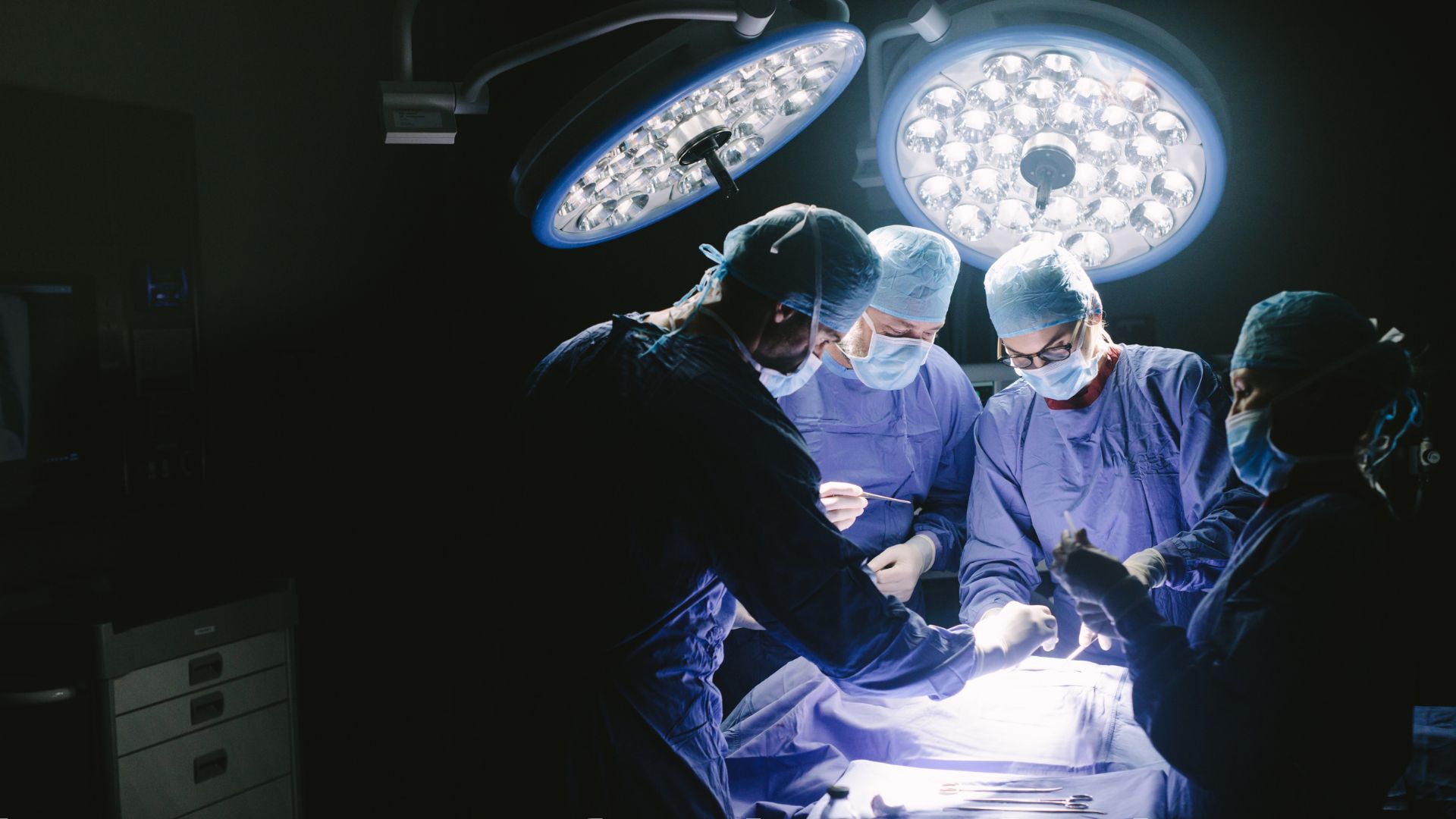 Four surgeons standing over a patient in an operating room with large powerful lights overhead. The surgeons are wearing blue scrubs, gloves, surgical masks, and hair nets.